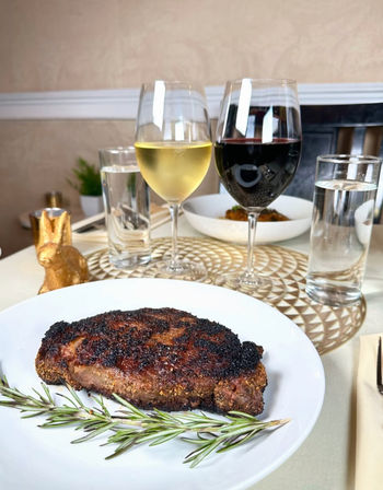 Pepper‑crusted steak on a white plate with rosemary sprigs, set on a restaurant table with red and white wine glasses, water tumblers and a small gold rabbit decor — elegant steak dinner and wine pairing.