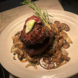 Seared steak medallions stacked on sautéed mushrooms and caramelized onions, garnished with a rosemary sprig and sliced scallion on a white dinner plate