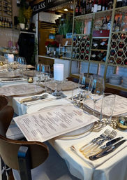 Elegant long dining table set for a private event in a fine-dining restaurant, with white tablecloths, printed menus, chargers, wine and water glasses, pearl-handled flatware, glass pillar candle holders, and a decorative wine rack backdrop.