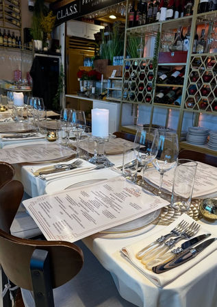 Elegant long dining table set for a private event in a fine-dining restaurant, with white tablecloths, printed menus, chargers, wine and water glasses, pearl-handled flatware, glass pillar candle holders, and a decorative wine rack backdrop.