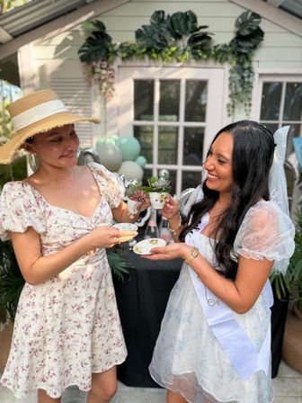 Two women in floral dresses at a backyard bridal-shower tea party, smiling and clinking teacups filled with succulents in front of a decorated garden shed.