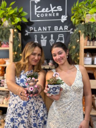 Two smiling people holding handmade succulent planters — a flower-crowned animal pot and a blue patterned pot — in an indoor plant bar with shelves of greenery.