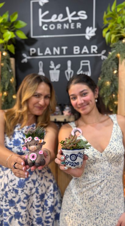 Two smiling people holding handmade succulent planters — a flower-crowned animal pot and a blue patterned pot — in an indoor plant bar with shelves of greenery.