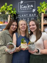 Three smiling women hold decorative succulent planters — a floral teacup, a smiling pot with heart-shaped glasses, and a stone face planter — inside a cozy indoor plant bar with shelves, green foliage and string lights.