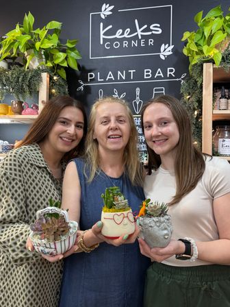 Three smiling women hold decorative succulent planters — a floral teacup, a smiling pot with heart-shaped glasses, and a stone face planter — inside a cozy indoor plant bar with shelves, green foliage and string lights.