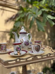 Charming vintage porcelain tea set with gold accents and illustrated cups and teapot arranged on a cream decorative serving tray in a sunlit garden