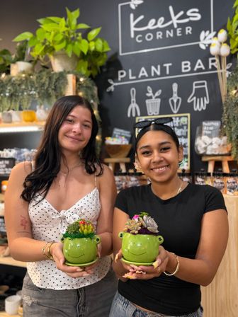 Two smiling friends holding green frog-shaped succulent planters at a cozy indoor plant bar, surrounded by hanging greenery, shelves of pots, and a chalkboard wall.