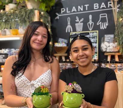 Two smiling friends holding green frog-shaped succulent planters at a cozy indoor plant bar, surrounded by hanging greenery, shelves of pots, and a chalkboard wall.