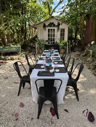 Inviting tropical garden dining scene: long white-tablecloth table with black metal chairs and place settings on a gravel courtyard, string lights and potted plants framing a small white cottage.