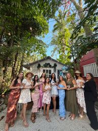 Group of smiling women in colorful dresses posing in front of a small white garden shed at a tropical backyard celebration, string lights overhead, lush palm foliage, and many holding small potted plants with blue star balloons nearby.