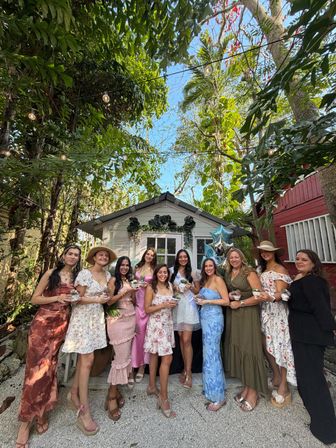 Group of smiling women in colorful dresses posing in front of a small white garden shed at a tropical backyard celebration, string lights overhead, lush palm foliage, and many holding small potted plants with blue star balloons nearby.