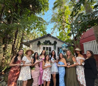 Group of smiling women in colorful dresses posing in front of a small white garden shed at a tropical backyard celebration, string lights overhead, lush palm foliage, and many holding small potted plants with blue star balloons nearby.