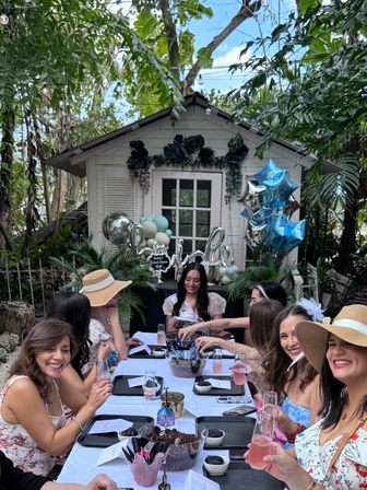 Lively tropical backyard garden party — women at a long white‑clothed table with pink champagne flutes, blue and silver star balloons, and a decorated white garden shed backdrop.