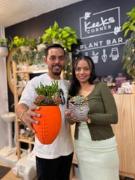 Two people holding succulent planters inside an indoor plant bar — a bright orange football-shaped planter and a decorative face-shaped concrete planter with succulents, surrounded by hanging greenery and wooden shelves.