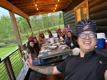 Person in a chef hat taking a selfie with a long table of friends wearing party crowns and bunny ears on a covered cabin porch, outdoor dinner with plates and string lights overlooking a green forest lawn.