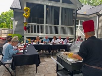 Outdoor backyard birthday on a suburban patio with guests seated at long black-tablecloth tables, a chef in a red hat cooking on a flat-top grill, and a gold '3' balloon by a screened porch.