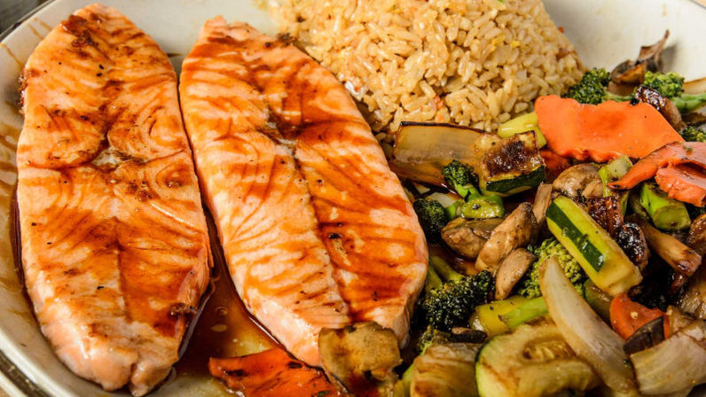Two glazed grilled salmon fillets with teriyaki-style sauce served with fried rice and colorful stir-fried vegetables (broccoli, zucchini, mushrooms, carrots) on a white plate