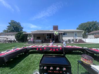 Sunny suburban backyard birthday party at a ranch-style house — U-shaped tables with black tablecloths and red decorations, kids with balloons on the covered patio, flat-top griddle in foreground.
