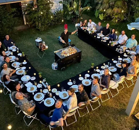 Overhead view of a backyard evening dinner party with U-shaped tables, guests in folding chairs, a chef in a red hat grilling on a large flat-top, plates and string lights in a lush garden.
