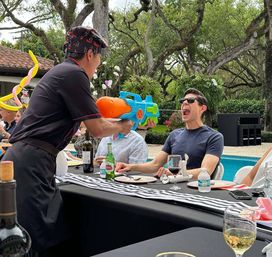Poolside backyard party scene with a server playfully squirting a large colorful water gun into a sunglasses-wearing guest’s open mouth during an outdoor luncheon by the pool, with drinks and balloon art on the table.