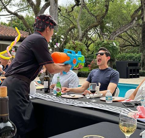 Poolside backyard party scene with a server playfully squirting a large colorful water gun into a sunglasses-wearing guest’s open mouth during an outdoor luncheon by the pool, with drinks and balloon art on the table.