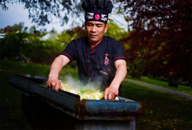 Outdoor chef in black uniform and tall hat cooking steaming colorful vegetables on a hot metal griddle in a leafy park setting