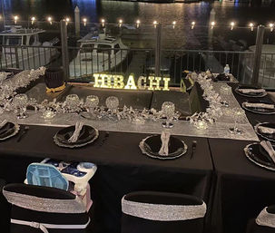 Nighttime waterfront hibachi setup with a lit HIBACHI sign, black-and-silver place settings and glittering centerpieces around the grill, string lights overhead and boats docked at the marina in the background.