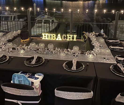 Nighttime waterfront hibachi setup with a lit HIBACHI sign, black-and-silver place settings and glittering centerpieces around the grill, string lights overhead and boats docked at the marina in the background.