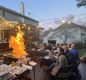 Evening suburban backyard deck dinner party with guests seated at a long table under string lights while a chef creates roaring grill flames, people raise drinks and cheer.