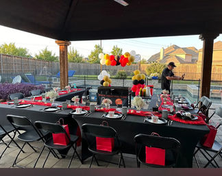 Festive backyard poolside party under a covered patio: long black-clothed banquet table with red runners, place settings and small floral centerpieces, red gift bags on folding chairs, red-black-yellow-white balloon arch, and a person cooking at a portable grill by the pool.
