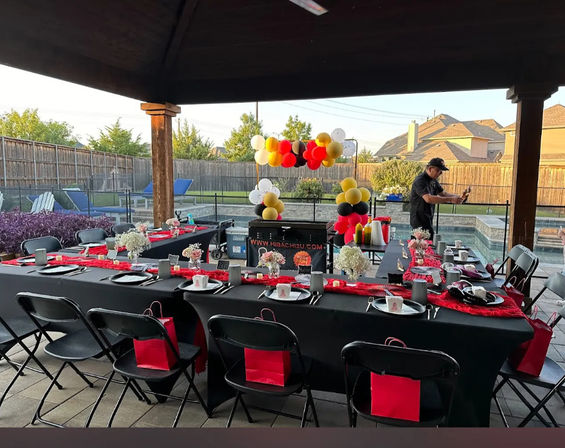 Festive backyard poolside party under a covered patio: long black-clothed banquet table with red runners, place settings and small floral centerpieces, red gift bags on folding chairs, red-black-yellow-white balloon arch, and a person cooking at a portable grill by the pool.