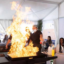 Masked teppanyaki chef flambéing on a large flat-top grill under a white event tent, towering flames as seated diners watch