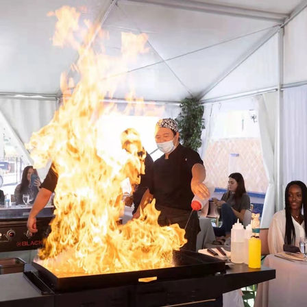Masked teppanyaki chef flambéing on a large flat-top grill under a white event tent, towering flames as seated diners watch