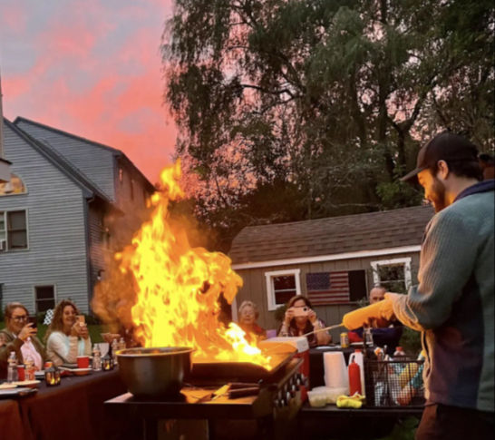Backyard sunset cookout with a chef flambéing a large grill, tall orange flames rising as guests watch and take photos in front of houses and trees.