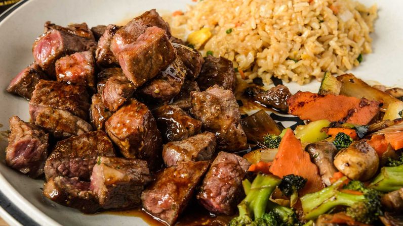 Close-up of bite-sized seared steak cubes glazed in savory sauce, served with fried rice and stir-fried broccoli, carrots, and mushrooms on a plate