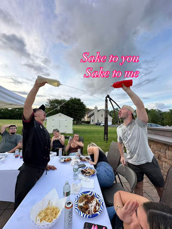 Backyard party scene: two people standing at a long outdoor table playfully squirt sauce from squeeze bottles into each other’s mouths in high-arching streams while friends laugh, plates of food and drinks on the table, lawn and houses beyond under a cloudy sky.