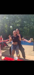 Two friends dancing at a sunny backyard pool party — a smiling woman in a black crop top and ripped jeans with a belly piercing is hugged from behind by a tattooed man kissing her cheek, picnic table with red tablecloth and condiments and wooded yard in the background.