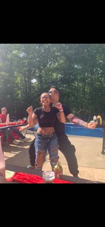 Two friends dancing at a sunny backyard pool party — a smiling woman in a black crop top and ripped jeans with a belly piercing is hugged from behind by a tattooed man kissing her cheek, picnic table with red tablecloth and condiments and wooded yard in the background.