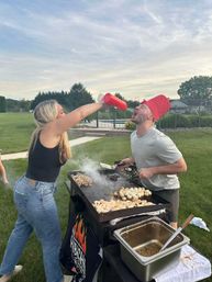 Playful backyard cookout: woman pours from a red condiment bottle into a man’s open mouth as he wears a red bucket hat while grilling chicken and potatoes on a flat-top griddle by a pool and green lawn at dusk.