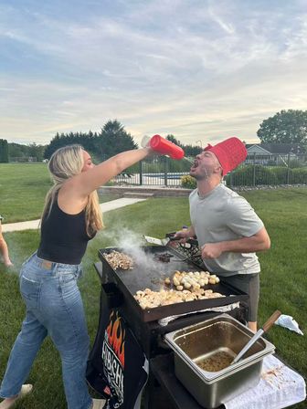 Playful backyard cookout: woman pours from a red condiment bottle into a man’s open mouth as he wears a red bucket hat while grilling chicken and potatoes on a flat-top griddle by a pool and green lawn at dusk.