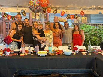 Group of smiling adults and a toddler posing under a "Happy Birthday" banner and balloons at a backyard patio party, long table with bowls, drinks, and string lights.