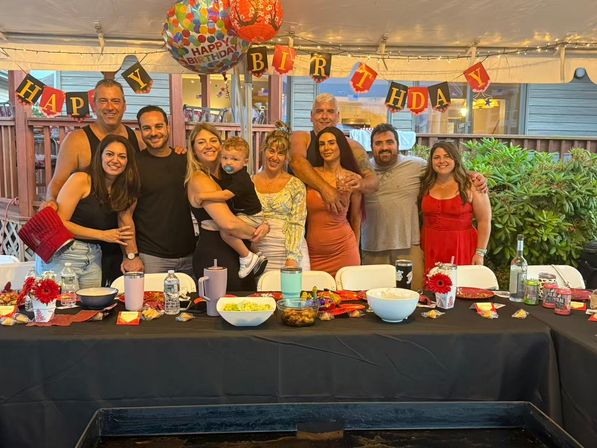Group of smiling adults and a toddler posing under a "Happy Birthday" banner and balloons at a backyard patio party, long table with bowls, drinks, and string lights.