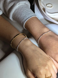 Close-up of two wrists wearing delicate silver chain bracelets and a gold bangle, resting on a white jewelry tray — minimalist stacked bracelet styling.