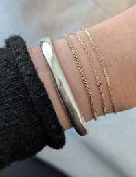 Close-up of a wrist with stacked bracelets: a chunky silver cuff and three delicate gold chains, one with a tiny pink gemstone, next to a black knit sleeve on a soft neutral background.
