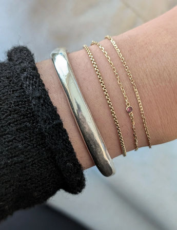 Close-up of a wrist with stacked bracelets: a chunky silver cuff and three delicate gold chains, one with a tiny pink gemstone, next to a black knit sleeve on a soft neutral background.