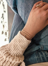 Close-up of a hand resting on denim knee, cream knit sweater sleeve, layered delicate gold chain bracelets with a tiny green gemstone and a gold floral ring.