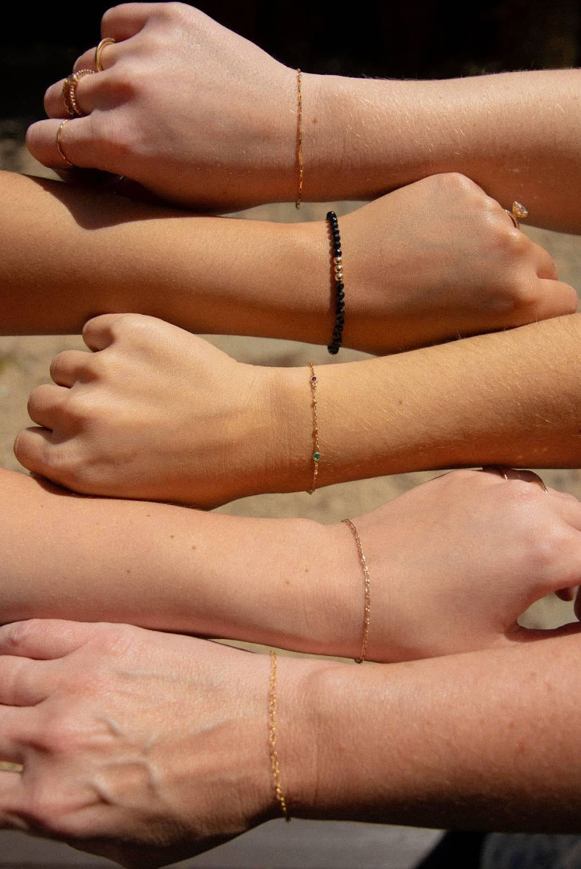 Sunlit close-up of five stacked wrists outdoors showcasing minimalist gold chain bracelets, a black beaded bracelet and simple rings — dainty, stylish jewelry on diverse skin tones.