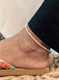Close-up of ankle with two anklets—a delicate gold chain and a small white pearl beaded anklet—paired with a floral-strap sandal on a woven mat, summer boho accessory shot.