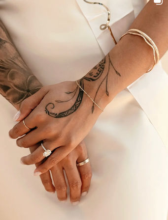 Close-up of tattooed hands over a white bridal dress, showcasing a sparkling diamond engagement ring, wedding band, delicate gold bracelets and a minimalist manicure.