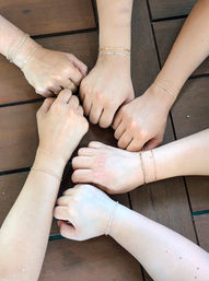 Six hands in a circle on a wooden table, each wrist wearing delicate gold and silver chain bracelets and a few minimalist rings — group bracelet styling shot